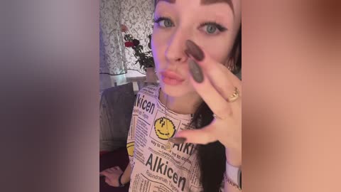 Video of a fair-skinned woman with blue eyes, wearing a black and white newspaper-patterned top. She has a subtle smile, green eyeshadow, and long, dark nails. Background includes a floral wallpaper and a vase with flowers.