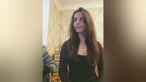 Video of a young woman with long brown hair, wearing a black top, standing indoors. Background features a beige floral wallpaper, a partially visible Scandinavian-style knit sweater, and a wooden dresser.