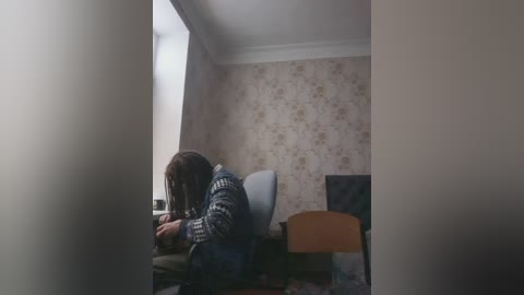 A video of a person in a small, dimly lit room with floral-patterned wallpaper, wearing a patterned sweater and sitting on a chair, looking down.