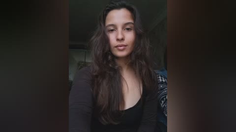 Video of a young woman with long, wavy dark hair, wearing a black top, standing indoors against a blurred background.