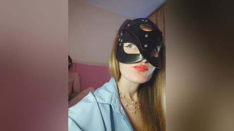 A video shows a woman with long blonde hair wearing a black leather mask and a light blue shirt, standing in a pink room with a person in the background.