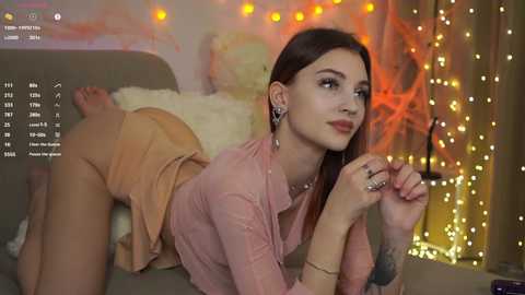 Video of a young woman with fair skin, brown hair, and small breasts, lying on a sofa, wearing a pink top and beige skirt. Background shows a cozy, warmly lit room with fairy lights.