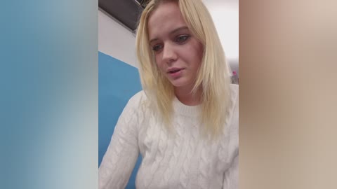 A video of a young blonde woman with fair skin and a slim build, wearing a white knitted sweater, looking down, in a blue and white room with a ceiling fan.