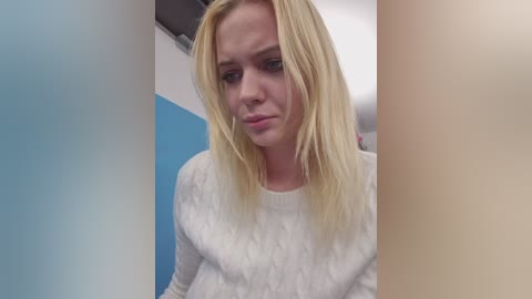 Video of a young, Caucasian woman with straight, shoulder-length blonde hair, wearing a white, textured sweater, in a room with blue and beige walls.
