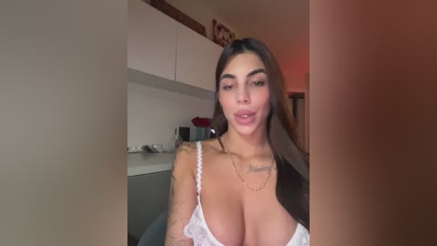 Video of a Latina woman with long dark hair, large breasts, and a tattoo, wearing a white lace bra, standing in a modern kitchen.