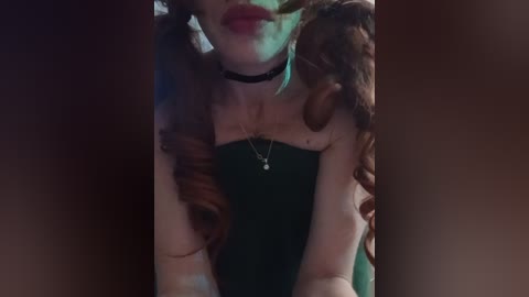 Video of a woman with fair skin, red hair, and a black choker, wearing a dark top, with hands intertwined around her, in a dimly lit room.