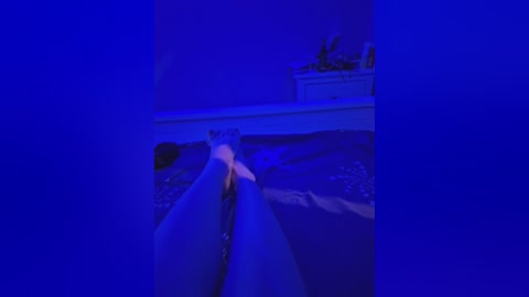 A video of a nude person, presumably female, with long dark hair, lying on their back on a dark surface, illuminated by blue light. The image is slightly blurred.