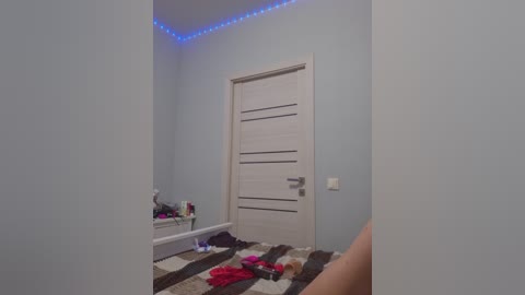 A video of a minimalist, light blue-painted bedroom with a white door and a striped rug. A person's legs are visible in the foreground, and a blue LED strip lights the ceiling.