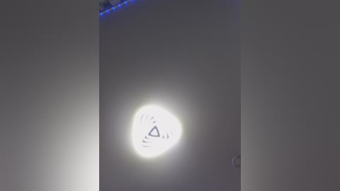 A video of a smartphone screen, with a white background, displaying a small, glowing, circular icon with a black triangle and dots, indicating a notification. The screen has a faint blue LED light at the top right corner.
