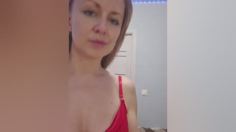 A video of a fair-skinned, light-haired woman in a red bra, taken indoors against a light blue wall with a white door in the background.
