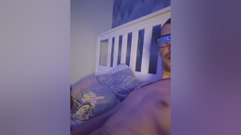 Video of a shirtless man lying in bed, wearing glasses, blue-patterned pajama pants, and a white pillow, with a blue wall and a white wooden headboard in the background.
