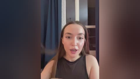 A video of a light-skinned woman with long brown hair, wearing a black sleeveless top, standing in a dimly lit room with blue curtains and wooden door in the background.