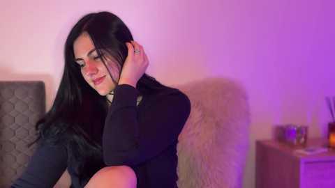 A video of a dark-haired woman in a black dress, with purple lighting, resting her head on a gray couch, looking serene.