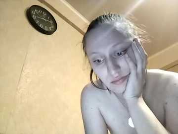 Video of a topless young woman with fair skin and wet hair, resting her head on her hand, standing in a bathroom with a beige wall and a round black clock.