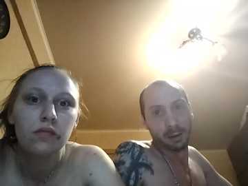 Video of a bald, shirtless man with a tattooed arm, standing next to a woman with fair skin and brown hair, both looking into the camera.