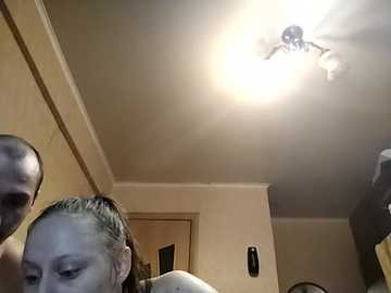 Video of a woman with short, light brown hair, wearing a white shirt, standing in a dimly lit room with beige walls and a ceiling fan.