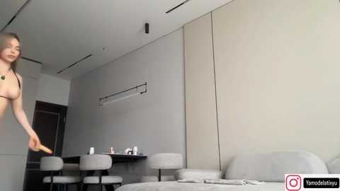 Video of a naked woman with small breasts, standing in a modern, minimalist bedroom with beige walls, a bed, and gray furniture.