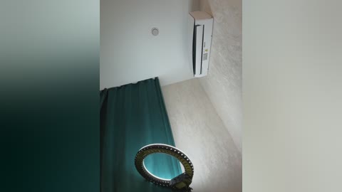 Video of a narrow, beige-walled room with a teal curtain partially covering a window. A circular, reflective object with a textured surface is prominently visible.