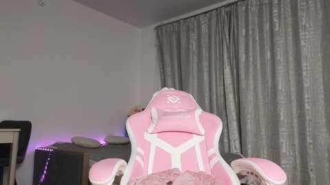 Video of a pink gaming chair with white accents, facing a gray curtain backdrop. A bed with beige pillows and a purple light strip is visible in the background.