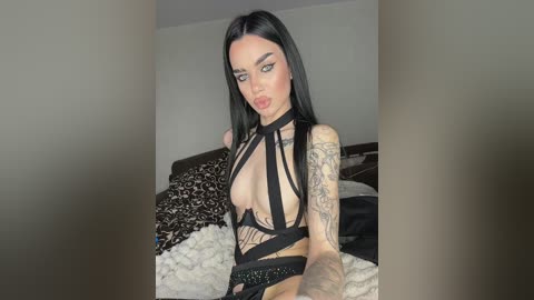 Video of a pale-skinned woman with long black hair and tattoos, wearing a black strappy top, sitting on a bed with a white blanket, in a dimly lit room.