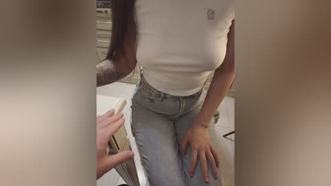 Video of a woman with medium-dark skin and long brown hair, wearing a tight white t-shirt and high-waisted light blue jeans, sitting on a white countertop.