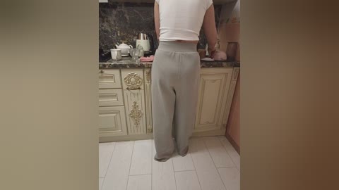 Video of a woman in a white top and grey high-waisted pants standing in a beige-tiled kitchen with white cabinets, granite countertops, and a black backsplash.