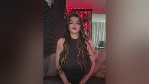 Video of a young woman with long, braided hair, wearing black makeup and a sleeveless top, seated in a dimly lit room with red LED lights, brick wall, and a bed in the background.