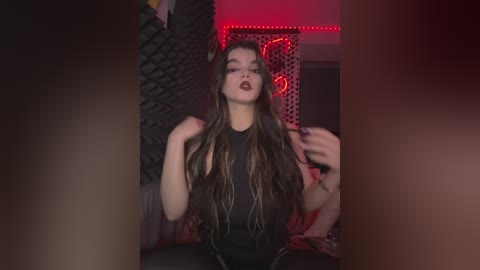 Video of a young woman with long, wavy brown hair, wearing dark makeup and a black top, sitting on a couch with a brick wall background, red LED strip lighting.