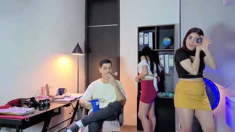 Video of a modern office with a white desk, lamp, and computer. Two Asian women, one in a yellow skirt, the other in a black top, take selfies.
