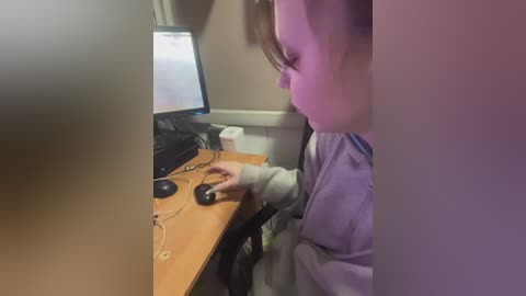 Video of a young child with light skin, wearing a gray hoodie, seated at a wooden desk, using a black computer mouse, with a monitor displaying a white screen.