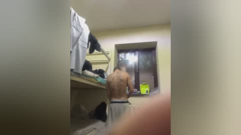 Video of a shirtless, muscular man with light skin and short brown hair, wearing white shorts, standing in a dimly lit, cluttered room with a bed, a window, and a neon green bottle.