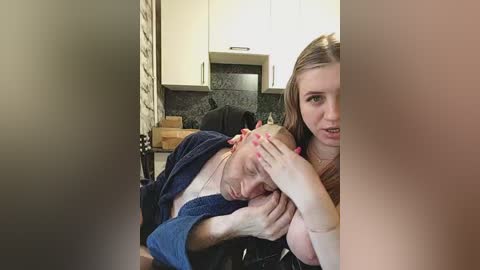 Video of a man in a dark robe cradling a bald woman in a light blue shirt, both with fair skin, in a modern kitchen with white cabinets and a dark countertop.