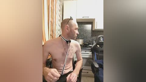 A shirtless, bald man in a black suit with a stethoscope around his neck sits in a kitchen chair, facing a gaming chair with headphones. The background features a modern kitchen with white cabinets, a black stove, and a gray backsplash.