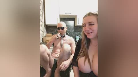 Video of a bald, muscular man with sunglasses and a black collar, sitting shirtless in a kitchen. Next to him, a young woman with large breasts and long hair, wearing a black top, smiles.