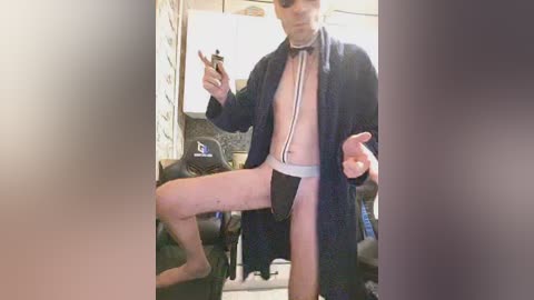 Video of a shirtless, slender man with light skin, wearing a black robe and a white diaper, posing playfully in a dimly lit room with a gaming chair and a partially visible laptop.