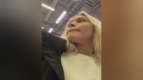 A video of a blonde woman with fair skin, wearing a black jacket over a white top and a silver choker, looking upward in a warehouse with fluorescent lights and exposed metal beams in the background.
