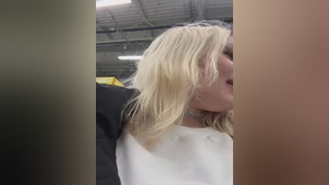 Video of a woman with light skin and blonde hair, wearing a white top, captured from the side. Background shows an industrial setting with metal beams and yellow equipment.