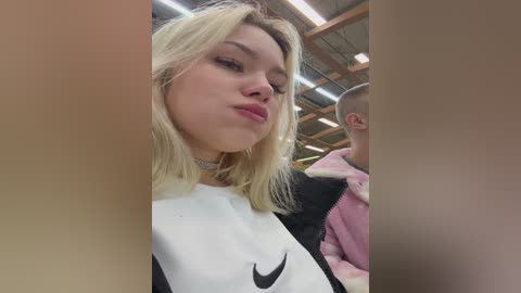 Video of a young blonde woman with a pouty face, wearing a black and white Nike sweatshirt, choker, and pink hoodie, standing indoors with a wooden ceiling and fluorescent lights.