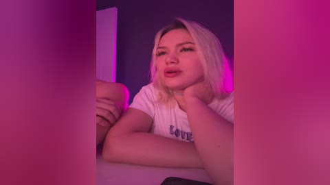 A video of a young woman with shoulder-length blonde hair, wearing a white t-shirt with \"LOVE\" printed in blue, sitting at a table with a pink and purple light backdrop.