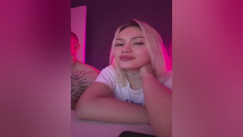 Video of a young Asian woman with long blonde hair, wearing a white T-shirt, looking at herself in a mirror. She has a tattoo on her left arm, and the room is dimly lit with pink and purple lights.