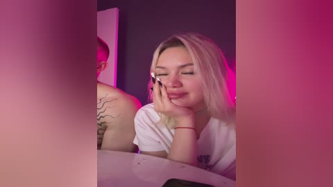 Video of a blonde woman with fair skin, small breasts, and medium build, applying makeup in a dimly lit room with purple and pink lighting. She wears a white t-shirt.