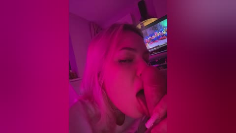 A video of a blonde woman with light skin, wearing a white top, licking an erect penis in a dimly lit room with purple and pink lighting, a TV displaying colorful images in the background.