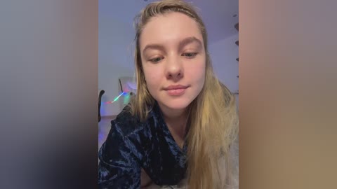 A video of a young Caucasian woman with long blonde hair, light skin, and closed eyes, wearing a dark velvet shirt, taken indoors with blurred background.
