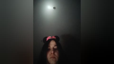 A dimly lit, grainy video of a woman with long black hair wearing a red and white checkered headband, eyes closed, facing upwards towards a small light bulb in a dark, narrow corridor.