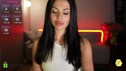 Video of a slender woman with long black hair and fair skin, wearing a white sleeveless top, standing indoors in a dimly lit room with a red glowing sofa and a blurry background.