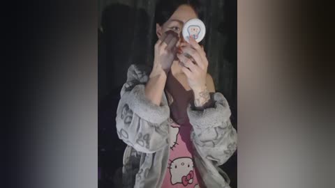 A video of a woman with dark hair, wearing a gray, fluffy jacket and a Hello Kitty t-shirt, taking a selfie with a phone. The background is dark and blurry.