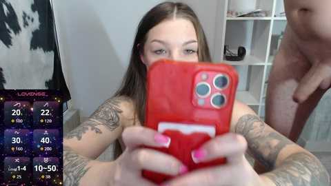 A video shows a young, pale-skinned woman with long brown hair, tattoos, and pink nails, holding a red iPhone, taking a selfie. The background features a cowhide patterned wall and a white shelving unit.