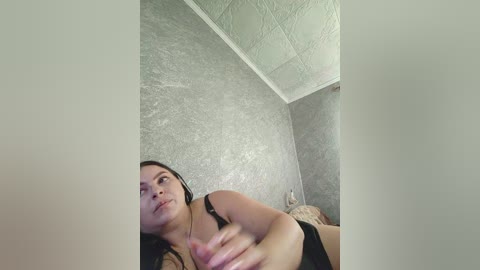 Video of a young woman with medium skin tone, dark hair, wearing a black bra, lying on a bed with textured gray walls and a ceiling with a patterned texture.