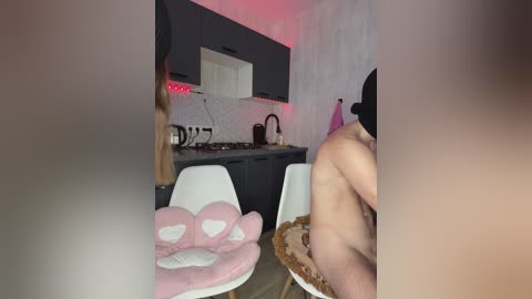 Video of a modern kitchen with a man in a black hat seated at a table, a pink stuffed animal chair in the foreground.