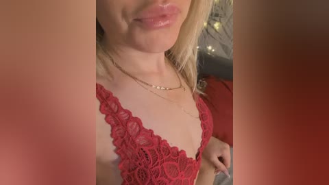 Video of a fair-skinned woman with blonde hair, wearing a red lace bralette, and a delicate gold necklace, with a blurred background featuring warm-toned fairy lights.